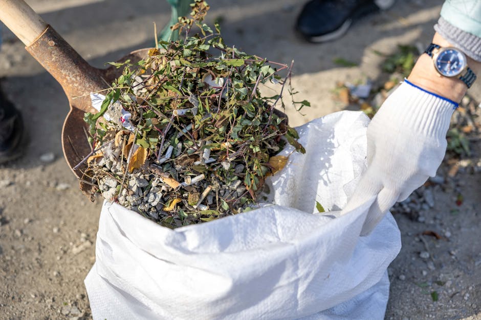A close-up view of a gardener's gloved hand holding a white gardening sack filled with a large pile of cuttings, including small branches, leaves, and some weeds. The gardener's wristwatch is visible 