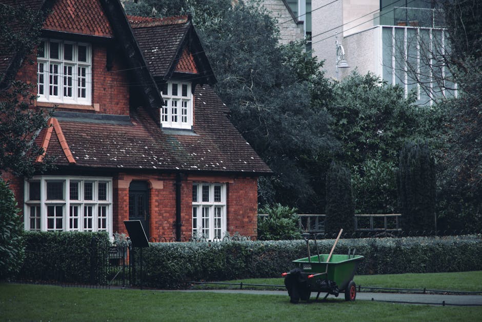 A red brick house with a steeply sloped, tiled roof featuring dormer windows and white-framed sash windows. The front garden has a well-maintained lawn bordered by a hedge, with a black metal fence al