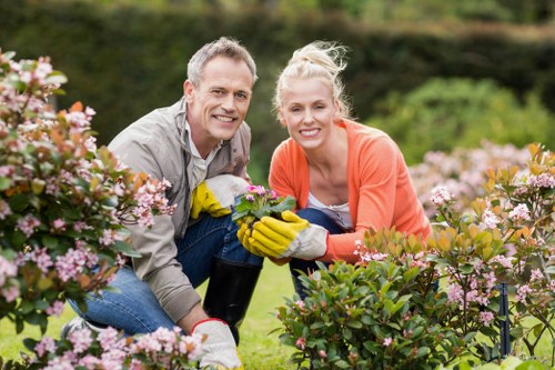 A man and a woman are kneeling together in a well-maintained garden, surrounded by pink and white flowering shrubs, with green foliage and grass in the background. The man has short, light-colored hair and is wearing a light grey shirt, while the woman has blonde hair tied back and is dressed in an orange top. Both are smiling and wearing yellow gardening gloves as they tend to a flowering shrub, suggesting active gardening or pruning work. The garden features a neatly arranged flower bed with various plants, lush green lawn areas, and a backdrop of trees and hedges, indicating a landscaped outdoor space. The image captures a sunny day, with natural light highlighting the vibrant colours of the flowers and foliage, reflecting the professionalism and care provided by a gardening service like Gardener Chessington. It emphasizes the benefits of outdoor maintenance, landscape enhancement, and garden nurturing.